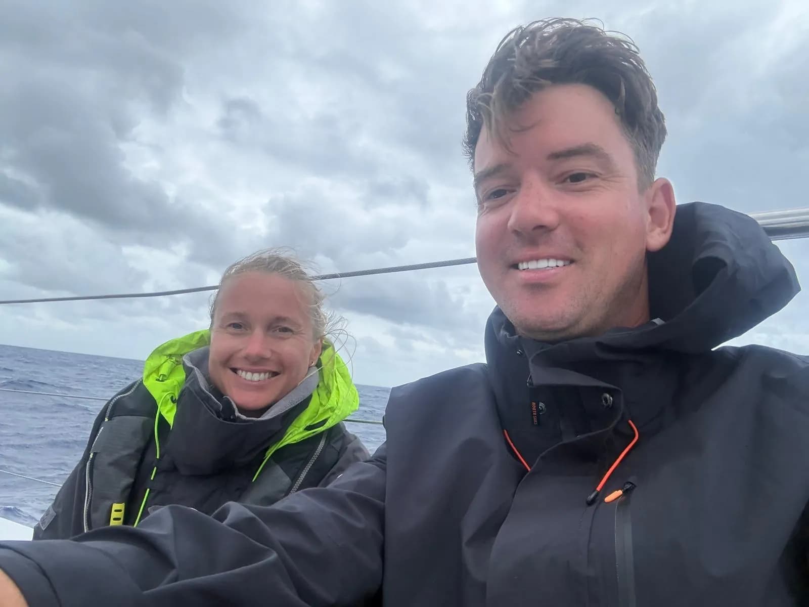 A man and a woman in sailing jackets smiling at the camera from a boat on a cloudy day.