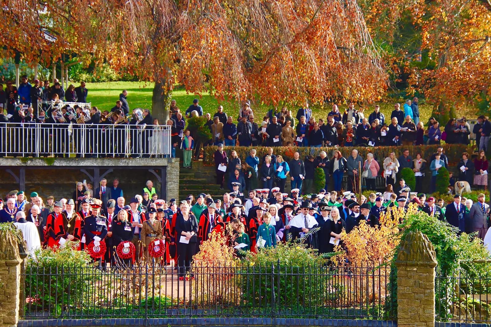 A large crowd of people, some in ceremonial dress and holding wreaths, gather outdoors under trees with autumn foliage.