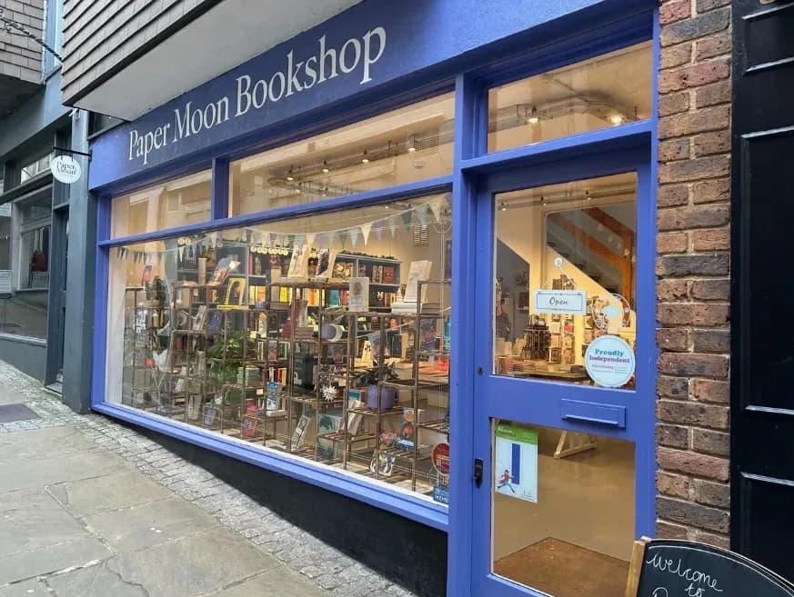 A purple storefront with a large display window for "Paper Moon Bookshop" showcasing books and various items.